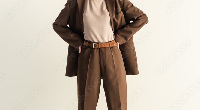 full length of young blonde woman in stylish brown suit standing with hands on hips on white